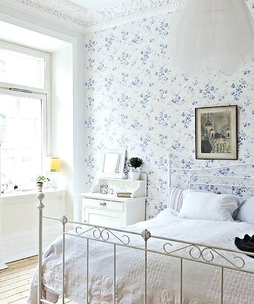 اتاق خواب با دیوار و تخت فلزی دونفره سفید رنگ که دیوار پشت تخت آن دارای کاغذ دیواری با طرح گل های ریز و زمینه سفید می باشد